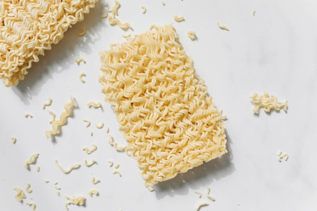 High-angle shot of raw instant noodles with scattered crumbs on a white background.
